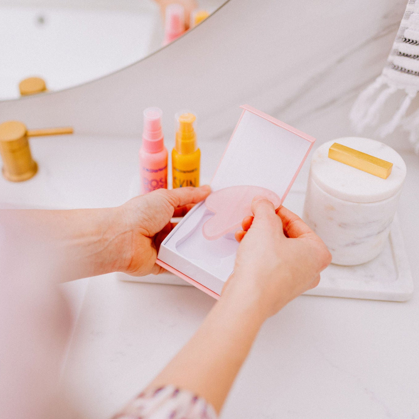 Frownies gua sha rose quartz stone being taken out of the box by a woman, the picture only shows her hands. Frownies Rose water hydrator spray and skin serum are also seen in the background of the picture