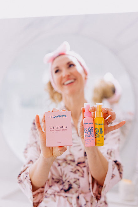 woman in background wearing pink skincare headband holding out pink box of gua sha, pink bottle of rose water hydrator, and yellow bottle of skin serum