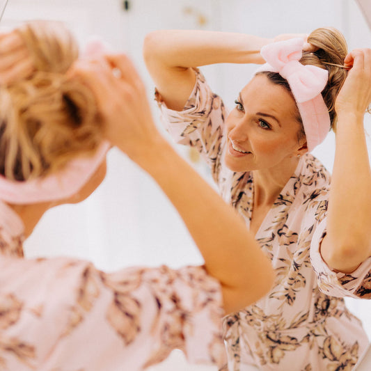 woman wearing a pink floral bathrobe and pink skincare headband with big bow adjusting her hair in the mirror