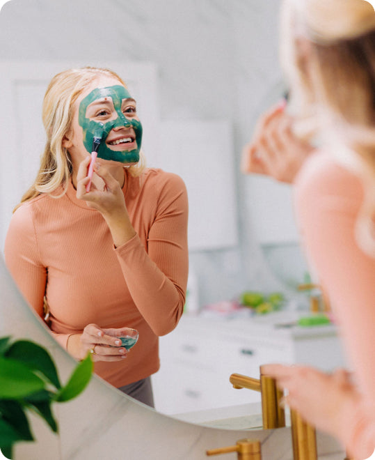 A blonde woman applies frownies green clay mud mask with applicator brush to face in front of a mirror