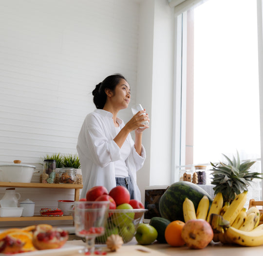 Woman drinking water with fruit and vegetables on counter.