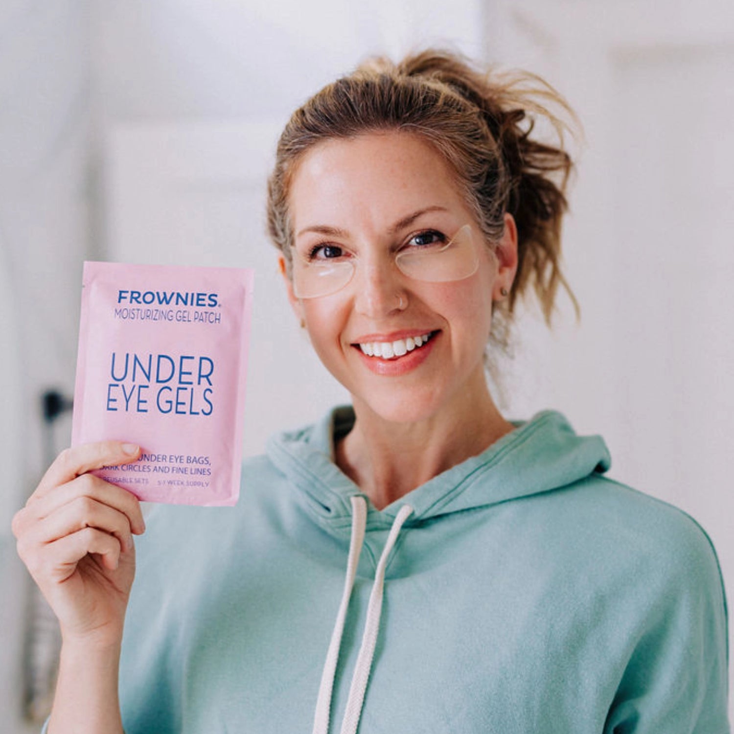 A smiling model holding the Frownies under eye gels package while wearing them as well