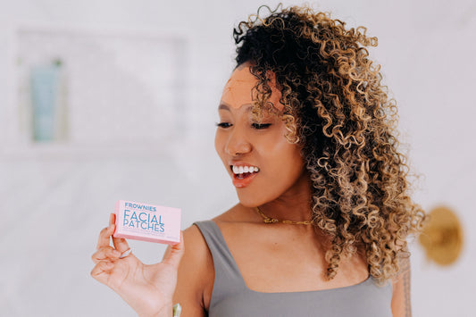 Woman smiling and holding a box of Frownies facial patches, with patches applied to her forehead