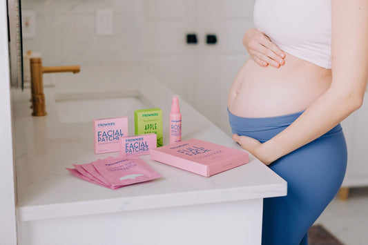 Woman with her hands placed on her pregnant belly next to a bathroom counter with a variety of Frownies skincare and Frownies facial patches