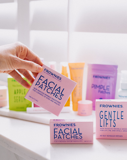 Hand holding a box of Facial Patches on a white surface with other Frownies products in the background.