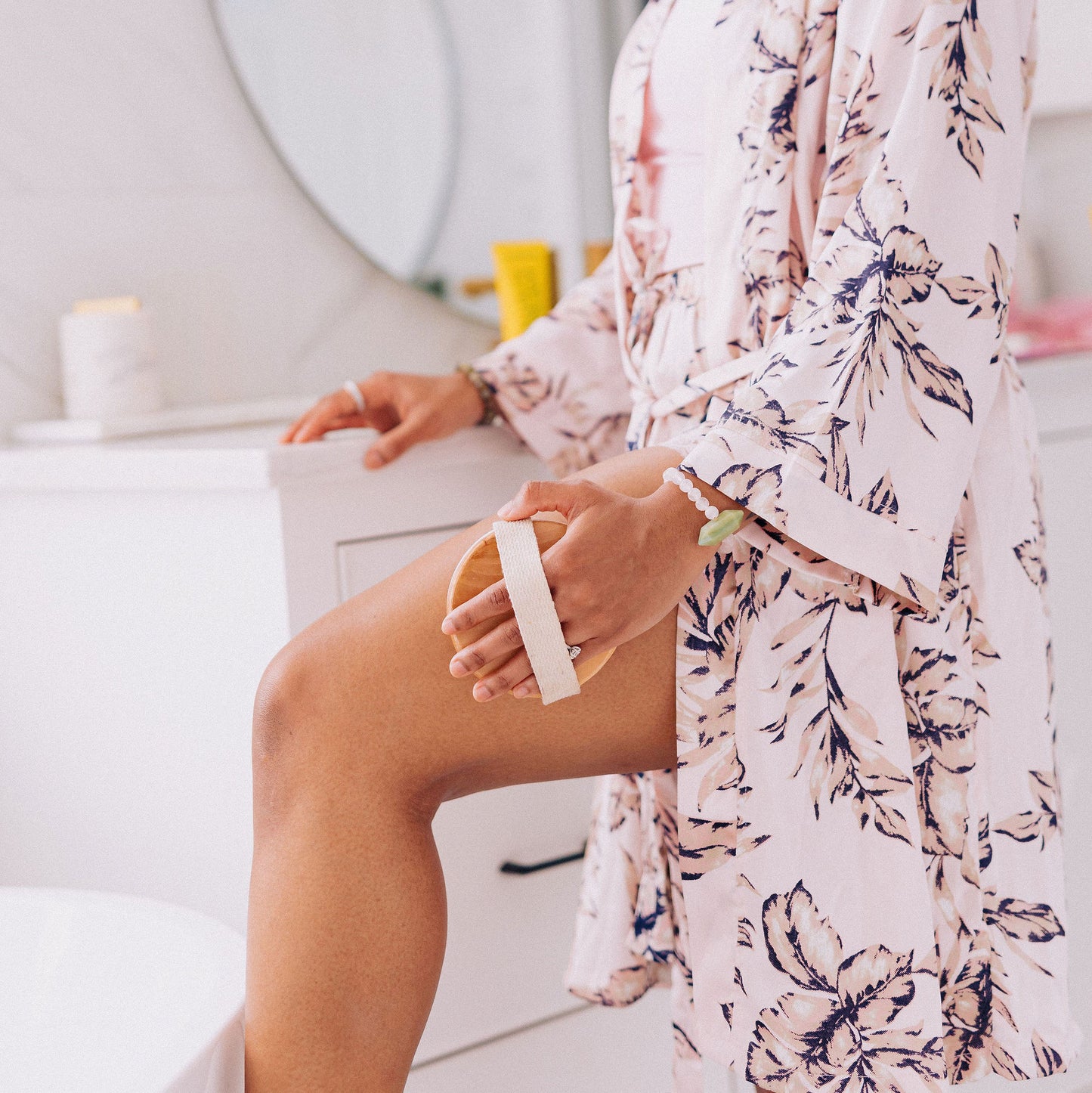 woman wearing pink floral bathrobe massaging thigh with natural boar's hair body brush for exfoliation and lymphatic drainage