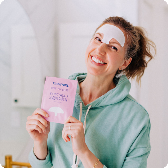 woman in light blue sweatshirt wearing white soft cotton forehead serum patch on forehead and holding pink package of frownies forehead serum patch