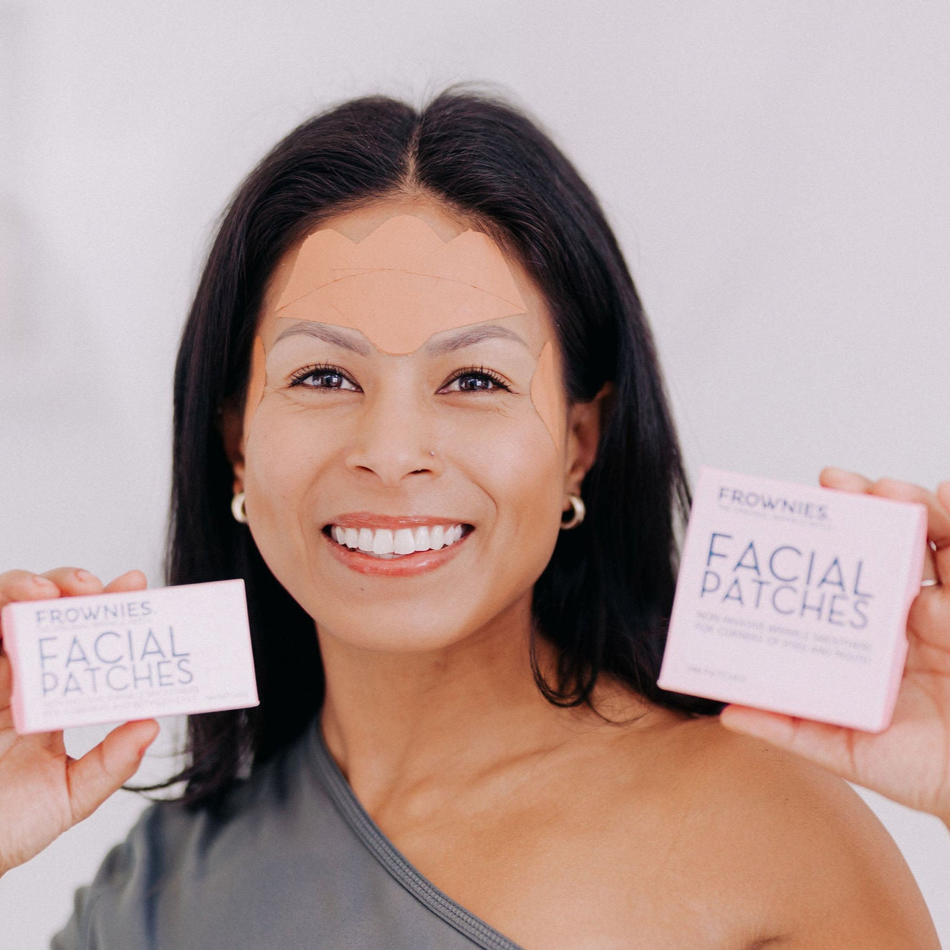 A woman with black hair shown smiling with both facial patches in her hand (FBE and CEM) while also wearing facial patches on her forehead area 