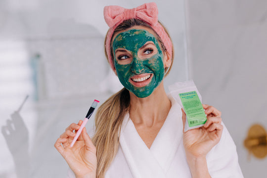 woman wearing pink skincare headband with bow holding skin care products green clay mask and applicator brush