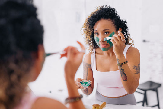 A woman applies face care clay mask with applicator brush in front of a mirror