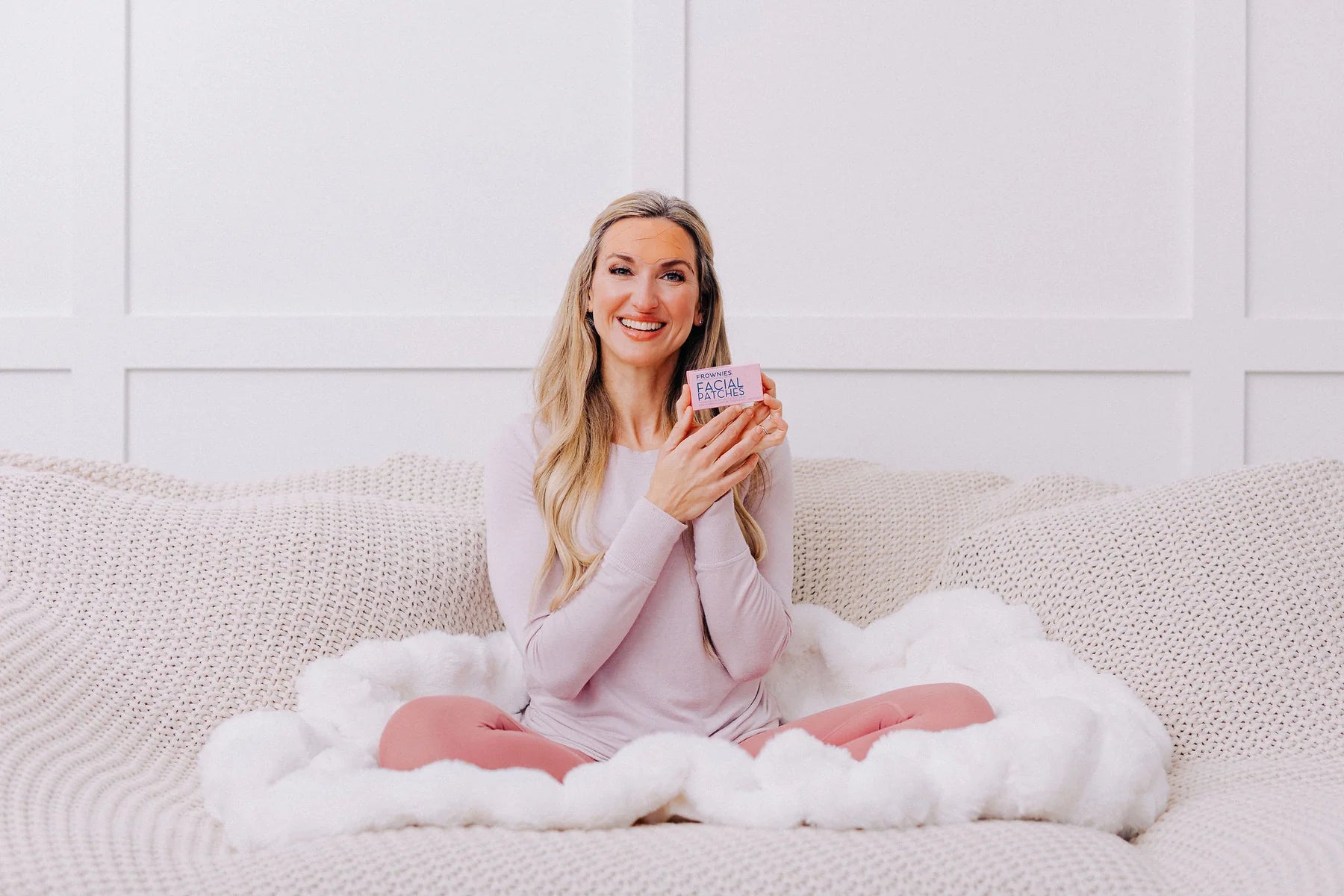 smiling woman with long blonde hair sitting on a couch with a fuzzy blanket holding a box of frownies facial patches for forehead and between the eyes