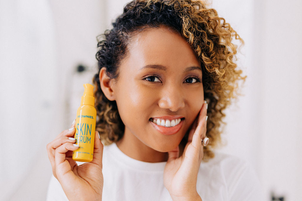 woman of color with curly hair touching face and holding yellow bottle of frownies vitamin C and E skin serum 