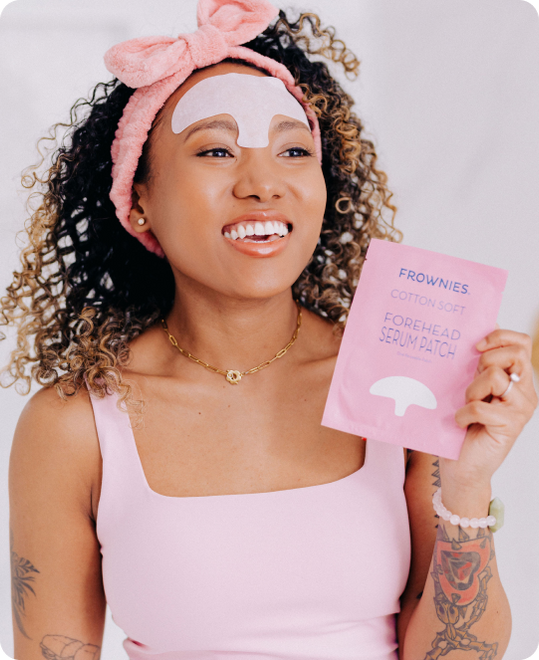 woman of color with curly hair and tattoos on arms wearing a pink skincare headband and wearing a white soft cotton forehead serum patch holding a pink package of frownies forehead serum patch