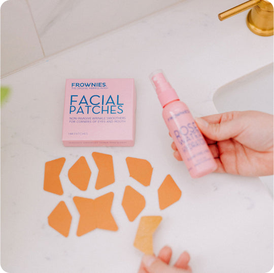 woman's hands holding pink bottle of frownies rose water hydrator with pink box of frownies facial patches for corners of the eyes and mouth and separated brown paper facial patches on countertop