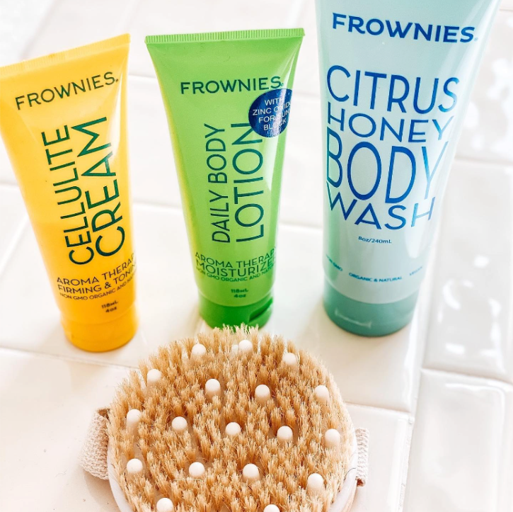 frownies cellulite cream, daily body lotion, citrus honey body wash, and round body brush on white bathroom tile