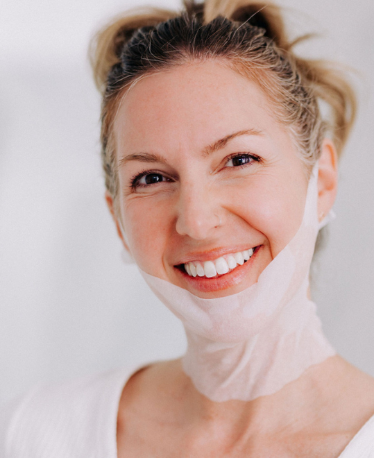 light skinned woman with hair pulled back smiling and wearing a frownies chin-up peptide mask over chin and jawline, down neck, and looped over ears