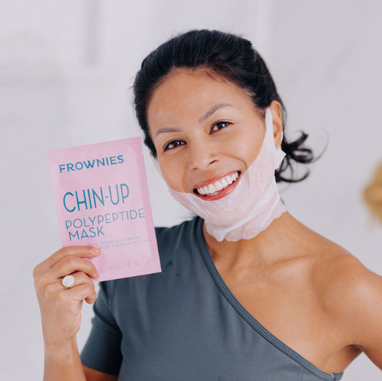 woman holding pink package of frownies chin-up polypeptide mask and wearing white soft cotton mask under lower lip, across neck and jawline and looped over ears