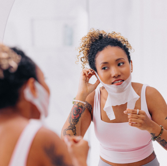 woman of color with curly hair pulled back and tattoos looking in mirror applying frownies chin up peptide mask over jawline and looped over ears