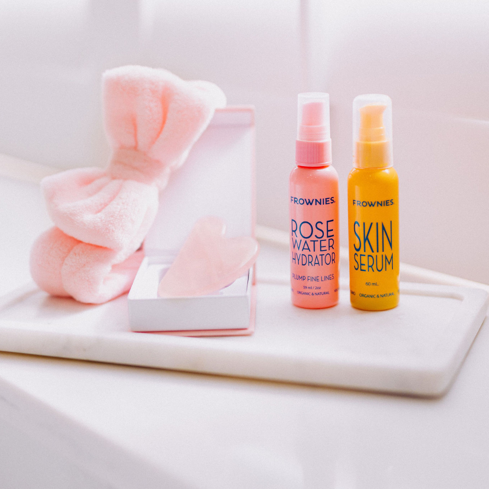 Frownies products including Rose water hydrator spray, skin serum, and Rose Quartz Gua Sha stone on a bathroom counter. 