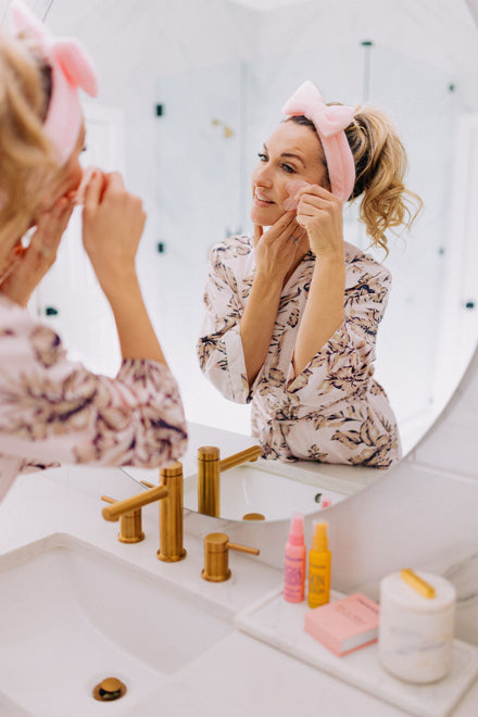 Woman using a gua sha stone in front of a mirror with various skincare items on the sink.