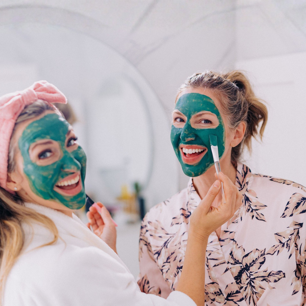 Two women applying green facial masks in a bathroom setting.