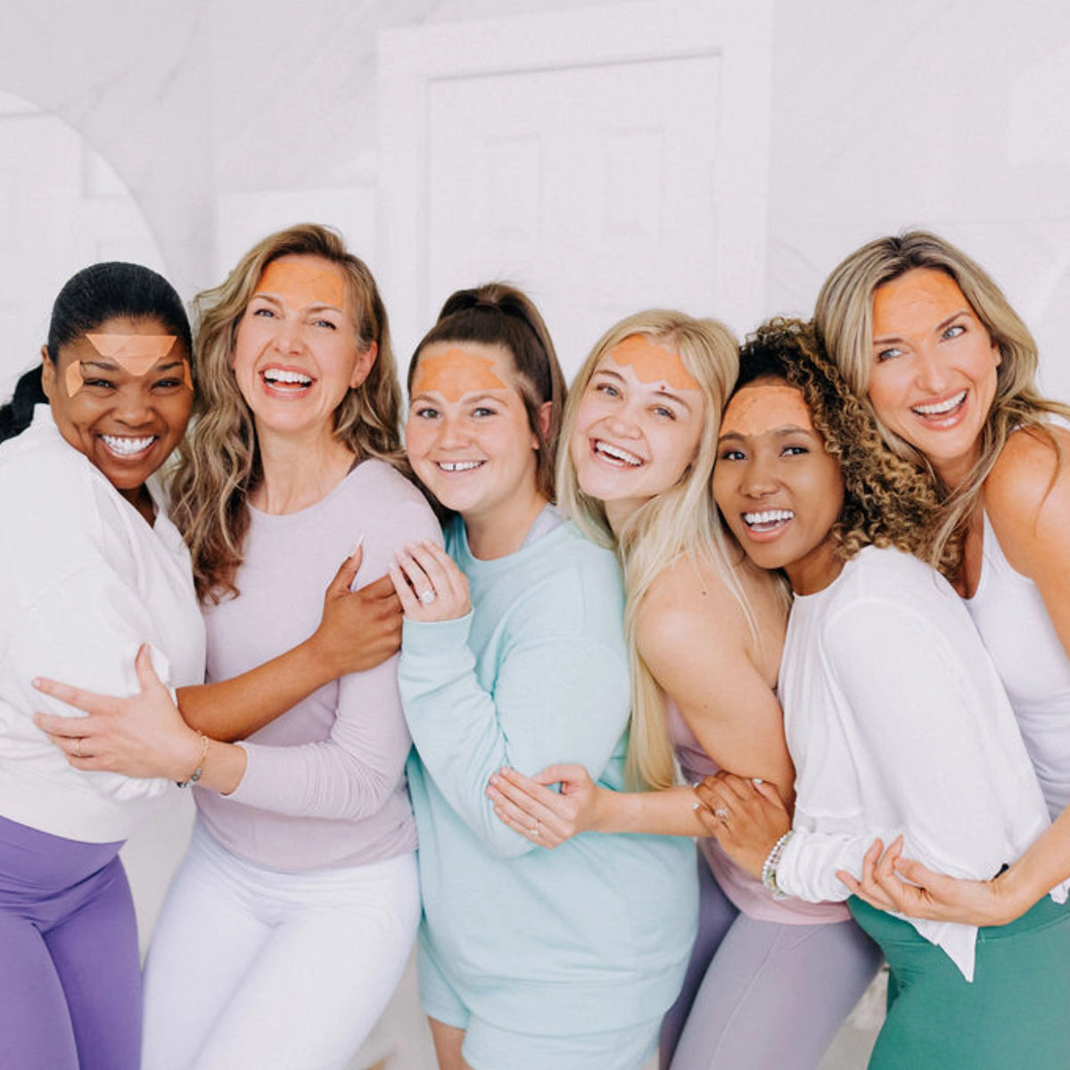 A group of six women close together, smiling and enjoying themselves while wearing Frownies facial patches.