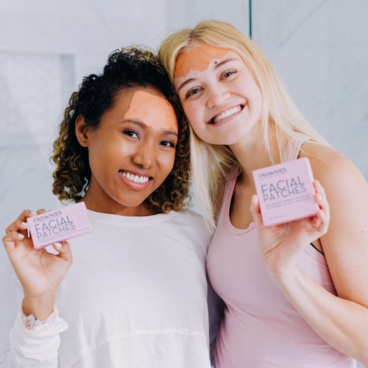 Two women smiling with their shoulders touching, wearing Frownies facial patches while holding the packaging up
