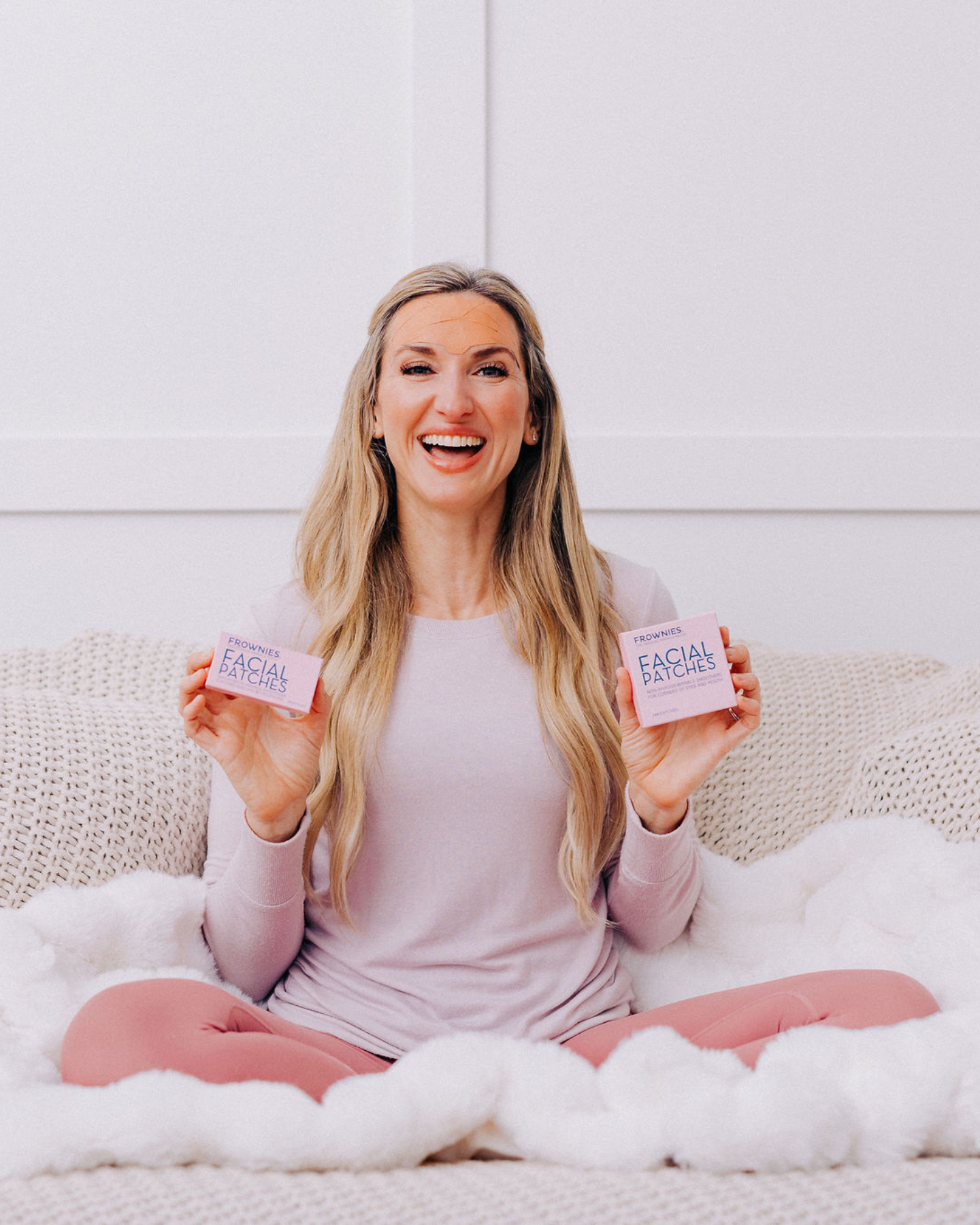 woman sitting on bed smiling holding two boxes of frownies facial patches for forehead and between the eyes and corners of the eyes and mouth