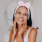 Woman applying a full face collagen mask to her face with a pink headband, smiling in front of a mirror.