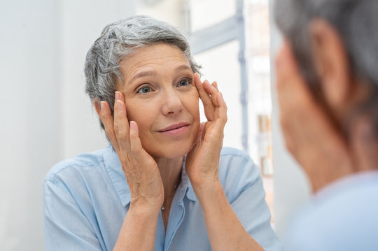 older woman with short gray hair touching her face and looking at her face in the mirror 
