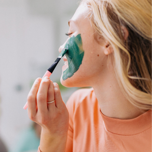 blonde woman applying Blue Green Algae Clay Mask with applicator brush to cheek