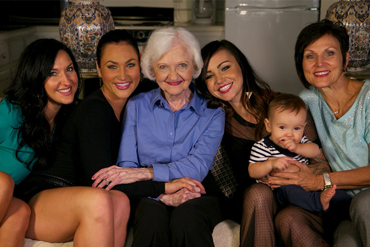 family photo of four generations of women including grandma with white hair in center surrounded by daughter and three adult granddaughters with dark hair and baby girl
