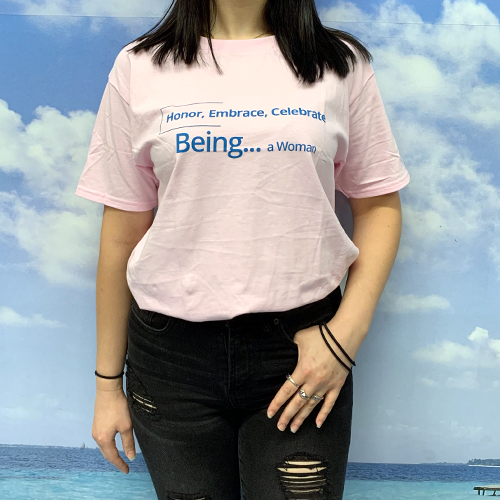Woman wearing a pink tee with blue text that reads Honor, Embrace, Celebrate, Being...a Woman
