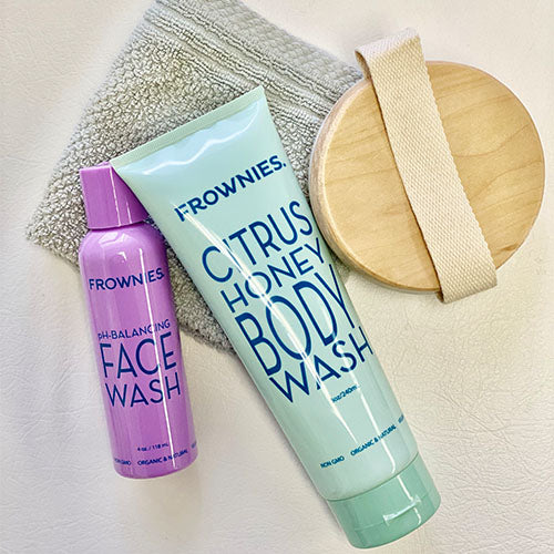  Frownies , pure natural preservatives,  ingredients matter, Frownies, pure and natural ingredients, Frownies light blue tube of Citrus Honey body wash  laying on a towel next to a body brush and a purple bottle of Frownies Face Wash